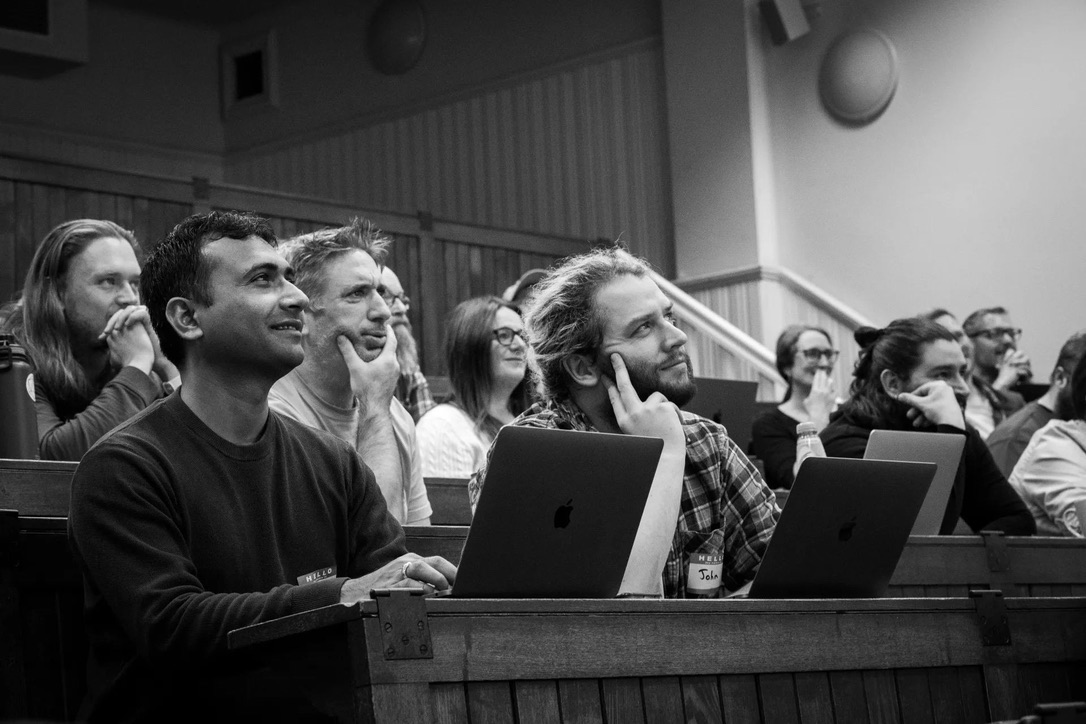 A black and white photograph of a room of Drupal developers and enthusiasts, with Sarsion in the forefront wearing a plaid shirt.